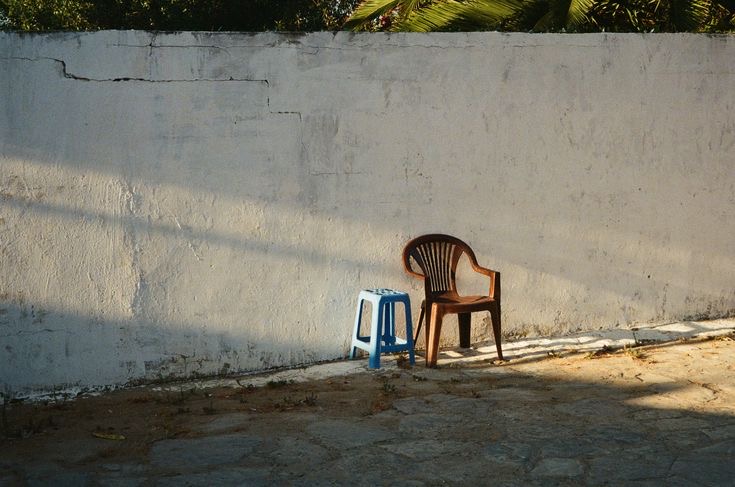 A wooden chair and a small stool standing apart against a sunlit wall.